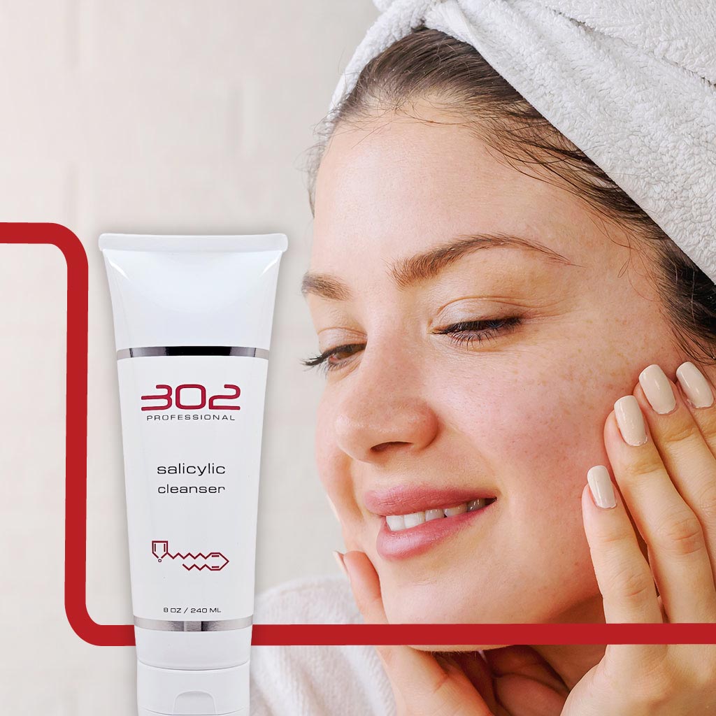SALICYLIC-CLEANSER-8OZ_WOMAN_2_1024x1024_72 | 302 Professional Skincare Close-up of a smiling woman with a towel on her head, applying skincare; white tube labeled '302 Professional Salicylic Cleanser' visible.