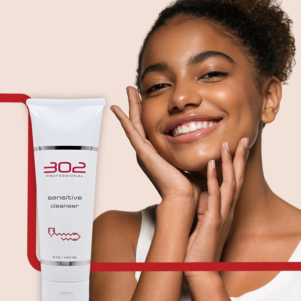 SENSITIVE-CLEANSER-8OZ_WOMAN_5_1024x1024_72 | 302 Professional Skincare Smiling woman with hands near her face beside a white tube labeled '302 Professional' Sensitive Cleanser, against a pale pink background.